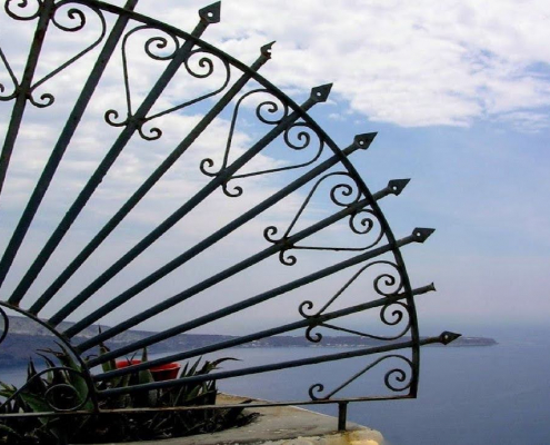 Das querformatige Foto zeigt eine Mittelmeerlandschaft. Vol der linken Fotoseite ragt ein Gitter aus Schmiedeeisen im Halbkreis in das Bild.