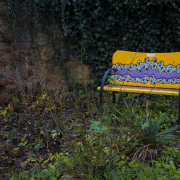 In einem Garten voller grüner Pflanzen steht eine gelbe Bank, die ein blaues Mosaik an der Rückenlehne hat.