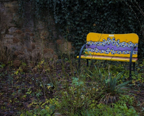 In einem Garten voller grüner Pflanzen steht eine gelbe Bank, die ein blaues Mosaik an der Rückenlehne hat.