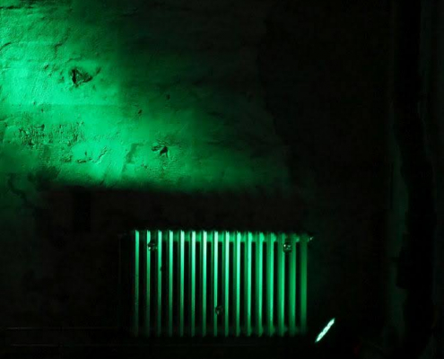 Auf dem hochformatigen Foto fällt ein grüner Lichtschein auf einen Zentralheizungskörper. Der restliche Raum ist dunkel.