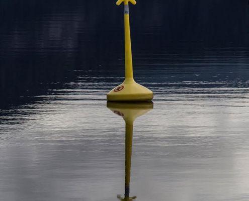 Ein hochformatiges Foto mit einer gelben Boje, die sich in dem blauschwarzen Wasser spiegelt.