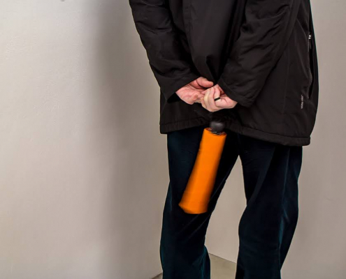2 weiße Wände eines Ausstellungsraums bilden eine Ecke vor der ein Mann mit Schwarzer Hose und schwarzer Jacke steht. Er ist von hinten zu sehen und hat einen orangefarbenen Schirm (Knirps) in der Hand.