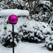In einem winterlichen Garten mit schneebedeckten Pflanzen und einem Vogelhäuschen ist eine matte lilafarbene Christbaumkugel befestigt.
