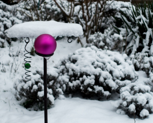 In einem winterlichen Garten mit schneebedeckten Pflanzen und einem Vogelhäuschen ist eine matte lilafarbene Christbaumkugel befestigt.