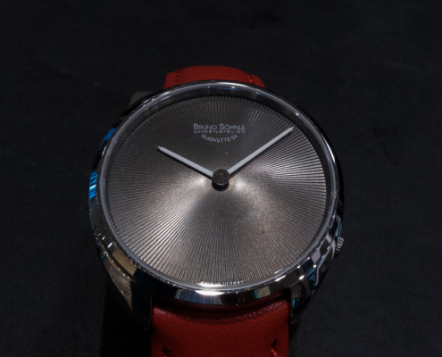 Das Ziffernblatt einer Bruno Söhnle Uhr mit rotem Armband auf schwarzem Hintergrund.