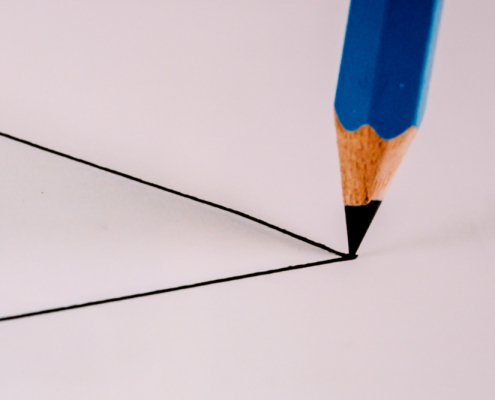 Von links ragen die Schenkel eines gezeichneten Dreiecks über die Mitte des Fotos. Auf die Spitze zeigt die Spitze eines blauen Bleistiftes.