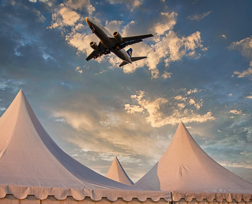 Über drei weißen Spitzen eines Zeltes fliegt ein Flugzeug