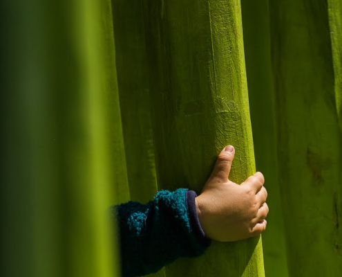 Auf dem hochformatigen Foto sind die Falten einen giftgrünen Vorhangs zu sehen. Eine Kinderhand, die aus einem türkisfarbenen Pullvover hervorschaut, greift eine Falte des Vorhangs.