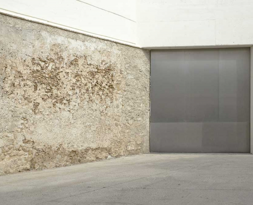 Ein weiß, grau, matschfarben gehaltenes Foto. Eine graue Stahltür grenzt an eine unverputzte, leicht erdfarbene Wand.