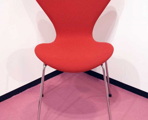 Ein roter Stuhl mit 4 Chrombeinen steht in der Ecke eines Raumes mit weißen Wänden, einem pinkfarbenen Teppich und einer schwarzen Scheuerleiste.