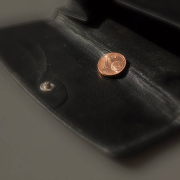 Auf einer aufgeklappten, schwarzen Geldbörse liegt eine 1 Cent Münze. Die Geldbörse liegt diagonal iBild auf einem grauen Untergrund.