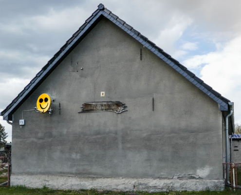 Das Foto im Querformat zeigt eine Seite eines Hauses mit einem Spitzdach. Die Wand dieser Seite ist grau. Eine Satelitenschüssel, auf der ein gelber Smiley formatfüllend aufgemalt ist, bildet den Lichtblick.el