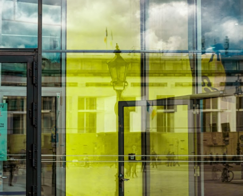 Das hochformatige Foto zeigt einen Teil der Glasfassade einen Geschäftshauses. In der Mitte dieser Fassade spiegelt sich eine gelbe Tür.