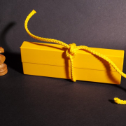 Auf dem Foto im Querformat steht in einer leichten Diagonalen ein länglicher, gelber Karton, der mit einer gelben Schnur zugebunden ist. An der linken Schmalseite des Kartons steht die Schachfigur Springer