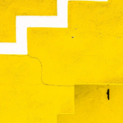 Das hochformatige Foto zeigt formatfüllend eine gelbe Wand. Am unteren Ende befindet sich eine gelbe Treppe. Von der linken Mitte des Fotos zur rechten oberen Ecke bildet eine weiße Zickzacklinie ein Muster. Von der Treppe zur weißen Zickzacklinie bildet ein Riss die Verbindung zwischen Treppe und Zickzacklinie.