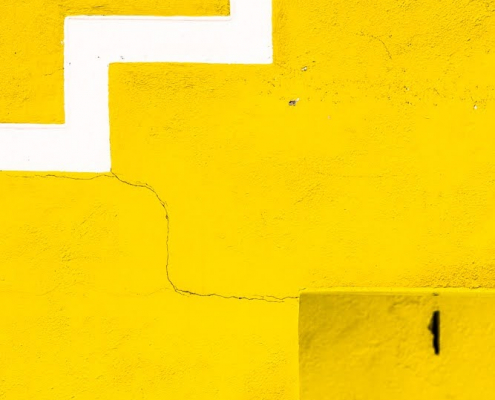 Das hochformatige Foto zeigt formatfüllend eine gelbe Wand. Am unteren Ende befindet sich eine gelbe Treppe. Von der linken Mitte des Fotos zur rechten oberen Ecke bildet eine weiße Zickzacklinie ein Muster. Von der Treppe zur weißen Zickzacklinie bildet ein Riss die Verbindung zwischen Treppe und Zickzacklinie.