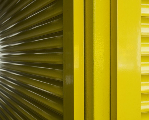 Auf dem Foto im Querformat sieht man zwei gelbe Lamellentüren, die an einem Pfosten, der in der Mitte des Bildes steht, befestigt sind.s Fotos