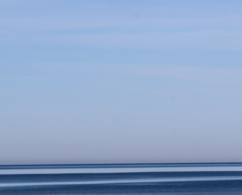 Das querformatige Foto zeigt zu 80 Prozent blauen Himmel und auf dem Rest blaues Wasser. Himmel und Wasser werden durch eine scharfe Horizontlinie getrennt. Das Wasser zeigt helle und dunkle parallel zum Bildrand verlaufende Linien.