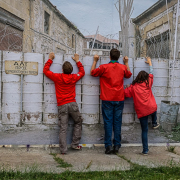 Das querformatige Foto zeigt den mit mannshoch gestapelten Fässern versperrten Durchgang zwischen zwei verlassenen Gebäuden. Drei Personen in roten Anoracks versuchen über die Absperrung zu schauen.