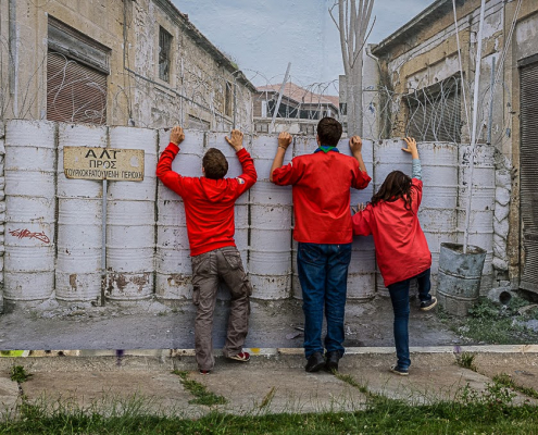 Das querformatige Foto zeigt den mit mannshoch gestapelten Fässern versperrten Durchgang zwischen zwei verlassenen Gebäuden. Drei Personen in roten Anoracks versuchen über die Absperrung zu schauen.
