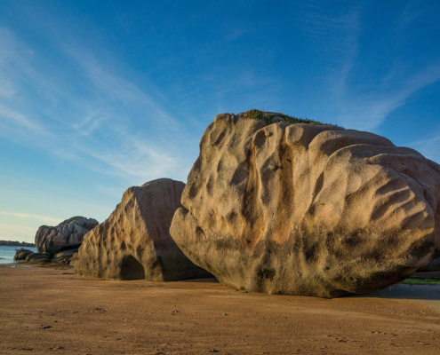 Das querformatige Foto zeigt drei große Granitblöcke an der Küste der Bretagne bei blauem Himmel.