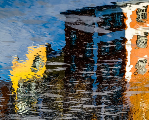 Das querformatige Foto zeigt ein orangefarbenes, ein dunkelrotes und ein gelbes Gebäude, die sich in bewegtem Wasser spiegeln und dadurch ein abstraktes Gemälde erzeugen.