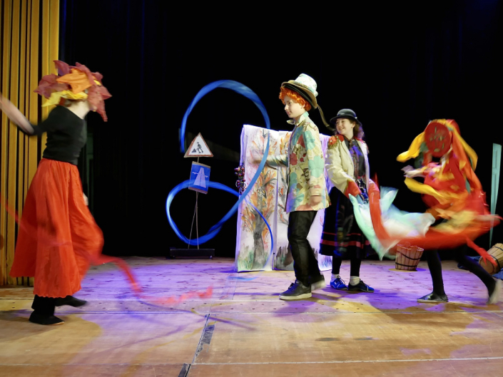 Vier Kinder auf einer Theaterbühne tanzen in Bunten Kostümen, Wasser gegen Feuer.