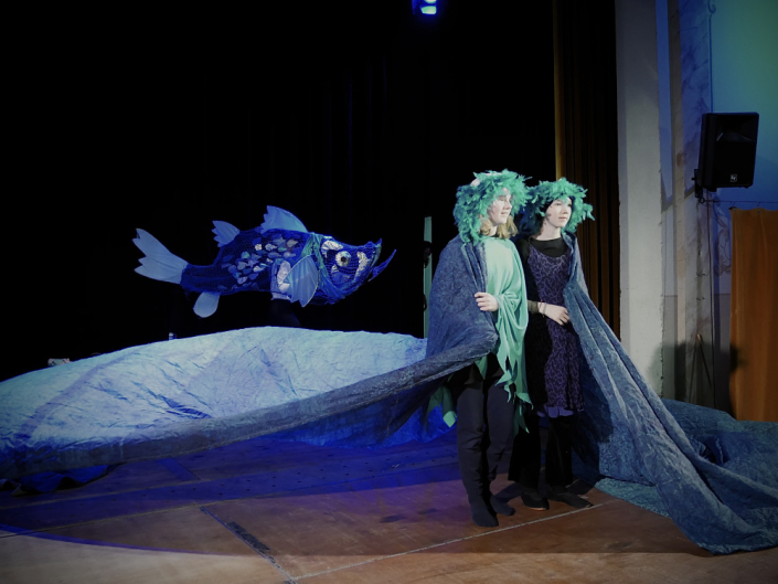 Zwei Mädchen auf einer Theaterbühne in langen Blauen Gewändern, im Hintergrund ein großer Blauer Fisch