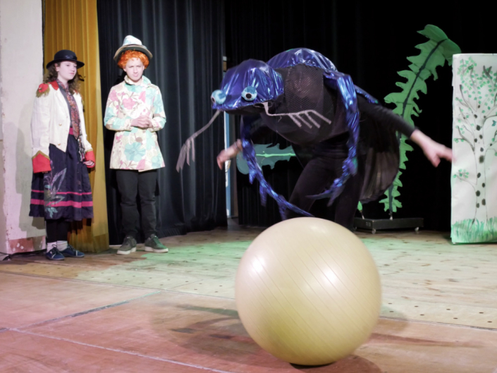 Drei Kinder auf einer Theaterbühne, eines in einem Skarabäus Kostüm rollt einen großen Ball