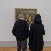 Auf dem querformatigen Foto steht ein älteres Paar vor einem Gemälde. Die Gesichter auf dem Gemälde schauen das Paar an.