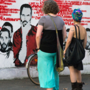 Auf dem hochformatigen Foto betrachten 2 junge Frauen die Parolen und Gesichter einer Graffitiwand.