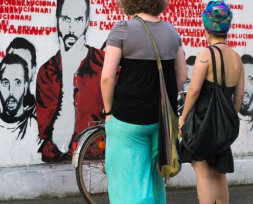 Auf dem hochformatigen Foto betrachten 2 junge Frauen die Parolen und Gesichter einer Graffitiwand.