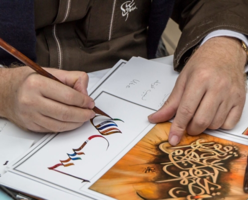 Das querformatige Bild zeigt die Hände eines Mannes, der in einer fremdem Schrift kalligraphiert.