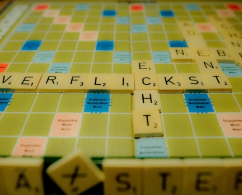 Das querformatige Bild zeigt den Ausschnitt eines Scrabble Spiels, auf dem das Wort 'Verflickst' im Blickpunkt steht. Ein schräg stehender Baustein mit einem X deutet auf den Schreibfehler hin.