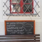 Das querformatige Bild zeigt das vergitterte Fenster eines Gasthauses. An dem Gitter hängt eine Schiefertafel mit dem Tagesgericht auf sächsisch.