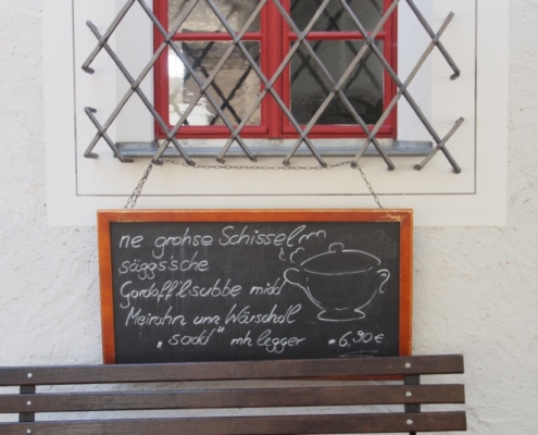 Das querformatige Bild zeigt das vergitterte Fenster eines Gasthauses. An dem Gitter hängt eine Schiefertafel mit dem Tagesgericht auf sächsisch.