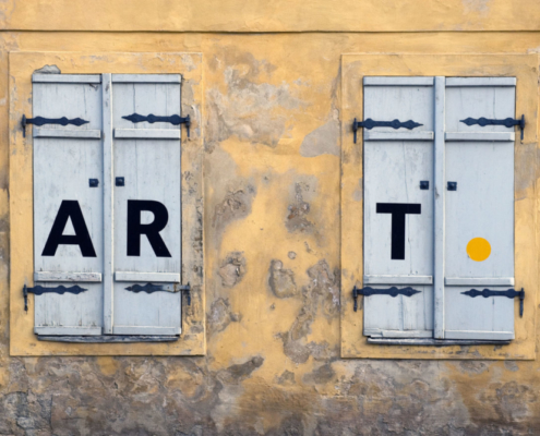 Das querformatige Bild zeigt eine abgeblätterte gelbe Wand mit 2 Fensterläden, auf denen die Buchstaben A R T prangen.