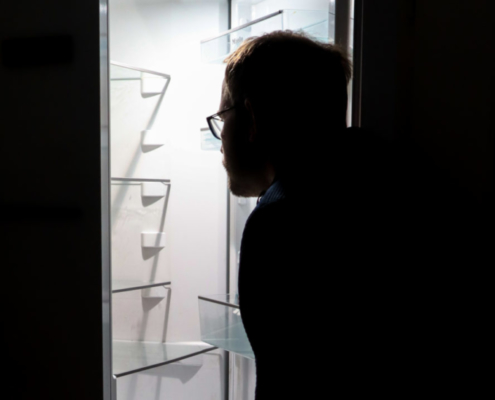 Das hochformatige Bild zeigt einen dunklen Raum, in dem ein Mann vor einem geöffneten, leeren Kühlschrank steht.