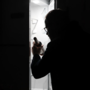 Das hochformatige Bild zeigt einen dunklen Raum, in dem ein Mann vor einem geöffneten, leeren Kühlschrank mit einer Flasche Bier in der Hand steht.
