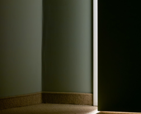 Das hochformatige Foto zeigt einen dunklen Raum, bei dem die Tür durch einen Spalt helles Licht aus dem Nachbarraum scheint.