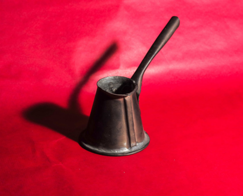 Das querformatige Foto zeigt ein einen kleine Kanne mit langem Stil auf rotem Hintergrund und den Schatten der Kanne.