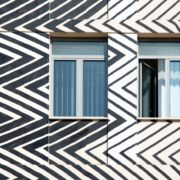 Das querformatige Foto zeigt einen Ausschnitt einer Hausfassade. Zu sehen sind zwei Fenster, die von schrägen dicken und dünnen Zackenlinien umgeben sind.