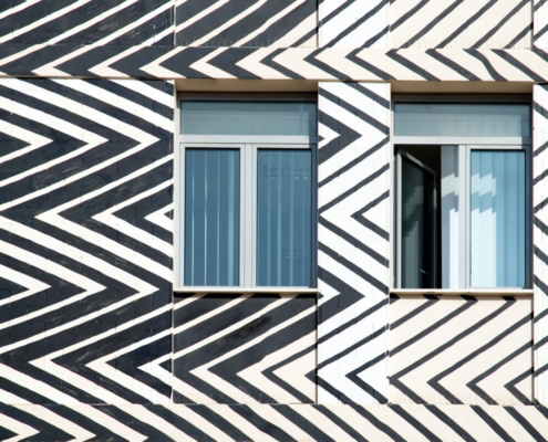 Das querformatige Foto zeigt einen Ausschnitt einer Hausfassade. Zu sehen sind zwei Fenster, die von schrägen dicken und dünnen Zackenlinien umgeben sind.