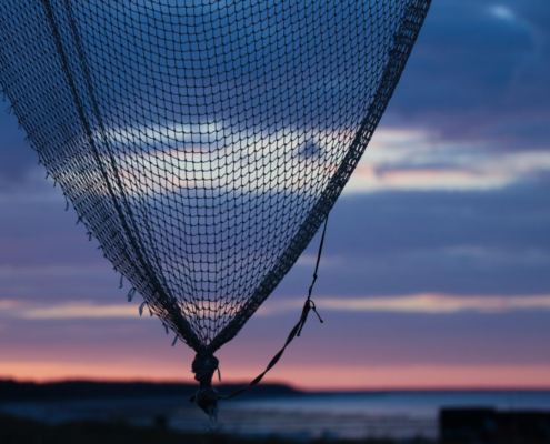 Auf dem querformatigen Foto blickt man im Vordergrund durch ein Fischernetz in den Himmel und im Hintergrund auf das Meer.