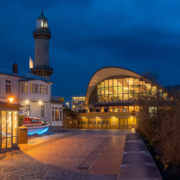 Das querformatige Foto zeigt die Strandhalle von Warnemünde mit dem Leuchtturm.