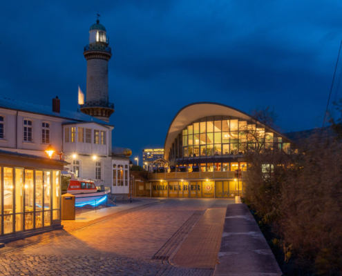 Das querformatige Foto zeigt die Strandhalle von Warnemünde mit dem Leuchtturm.