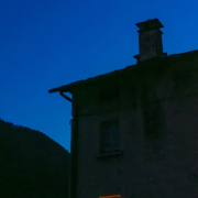 Das hochformatige Foto zeigt ein mehrstöckiges Haus im Gebirge mit einem im gelben Licht erleuchteten Fenster zur abendlichen blauen Stunde.