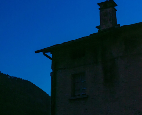 Das hochformatige Foto zeigt ein mehrstöckiges Haus im Gebirge mit einem im gelben Licht erleuchteten Fenster zur abendlichen blauen Stunde.