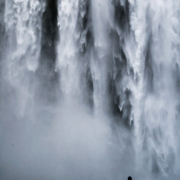 Das hochformatige schwarz-weiß Bild zeigt formatfüllend einen Wasserfall, der von oben runterfällt. Davor steht ein Mensch.