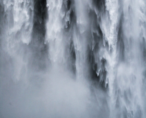 Das hochformatige schwarz-weiß Bild zeigt formatfüllend einen Wasserfall, der von oben runterfällt. Davor steht ein Mensch.